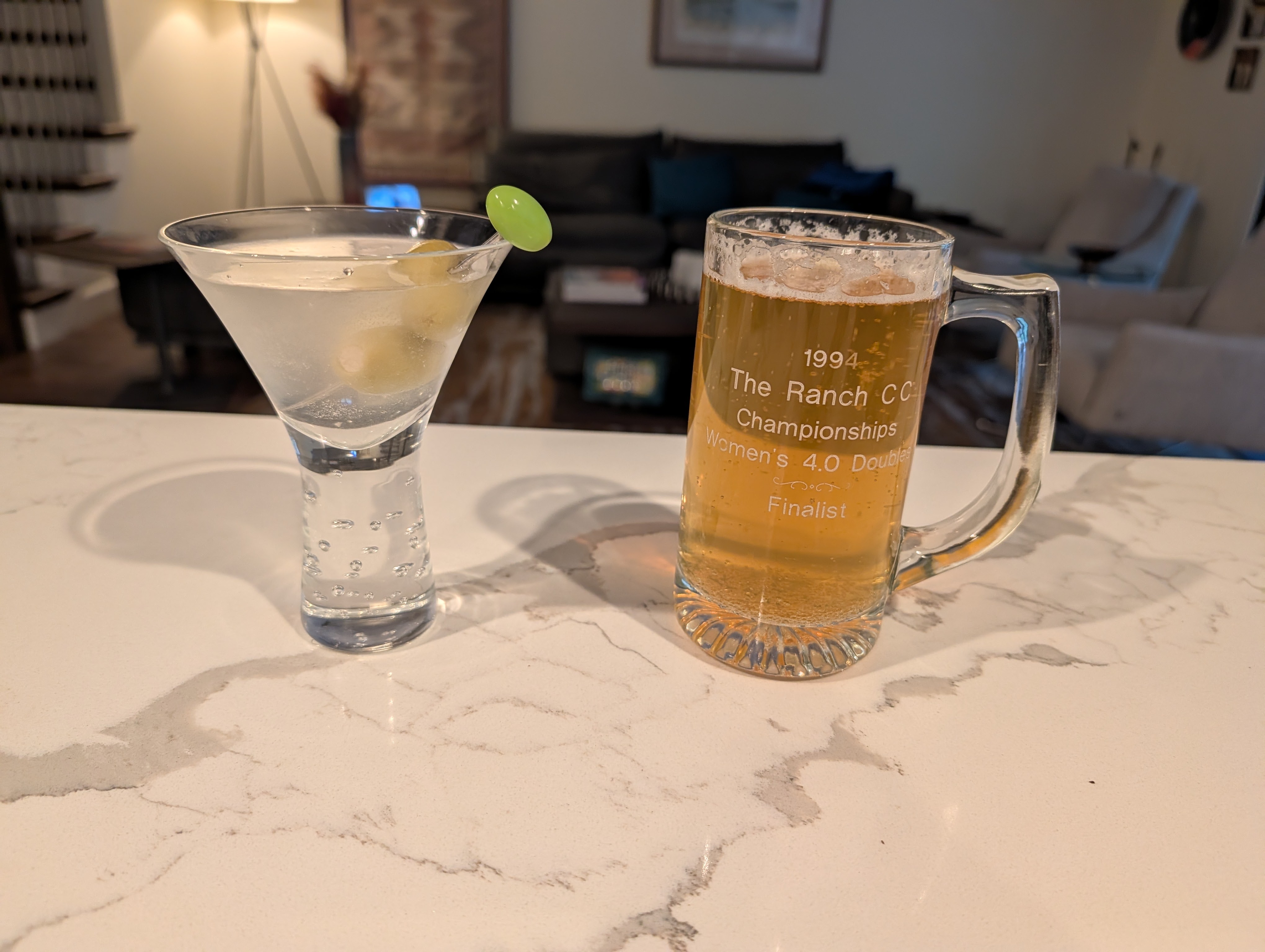 A martini glass with a clear cocktail and olives sits next to a large beer mug filled with golden beer, both on a light-colored marble countertop.