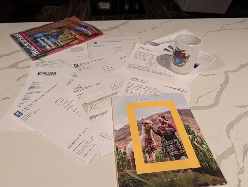 A table with a National Geographic Expeditions brochure featuring a woman and a llama, several invoices, and a mug.