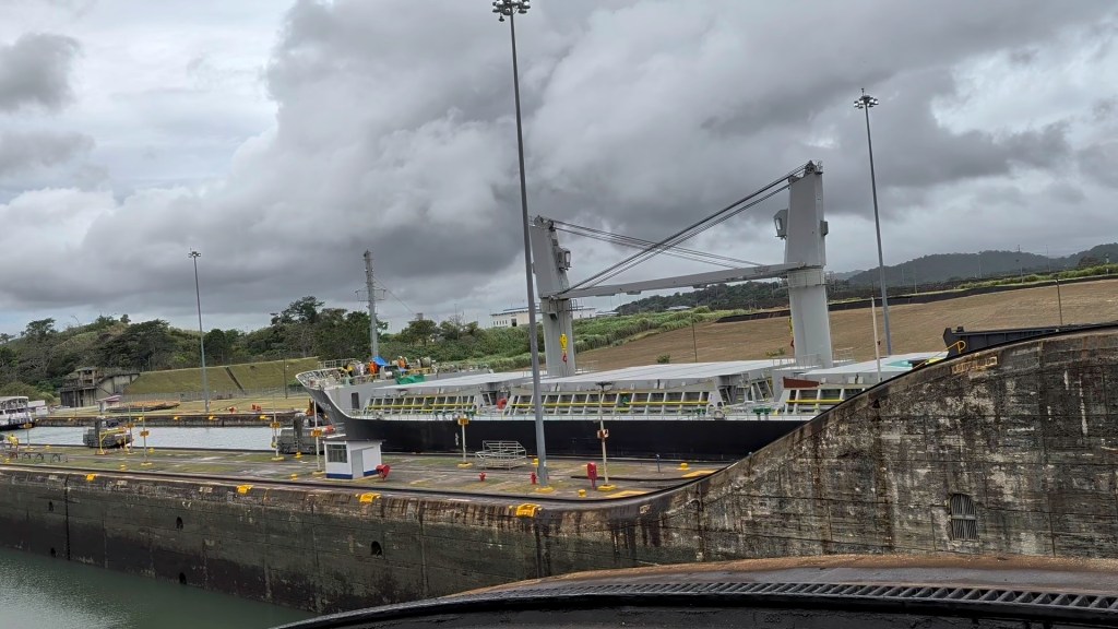 A large ship positioned in a lock chamber with cranes and machinery, surrounded by trees and cloudy skies.
