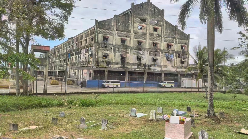 An abandoned, rundown building with broken windows and peeling paint, viewed from a grassy area with tombstones in the foreground and palm trees on the side.