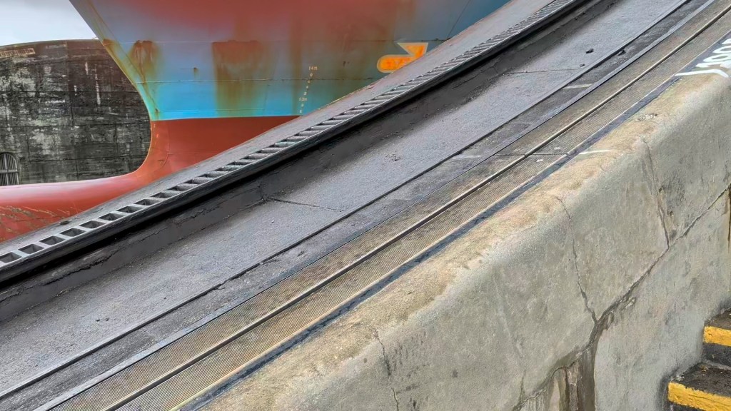 Close-up of a ship's hull partially visible behind a sloped dock, with textured surfaces and industrial details.