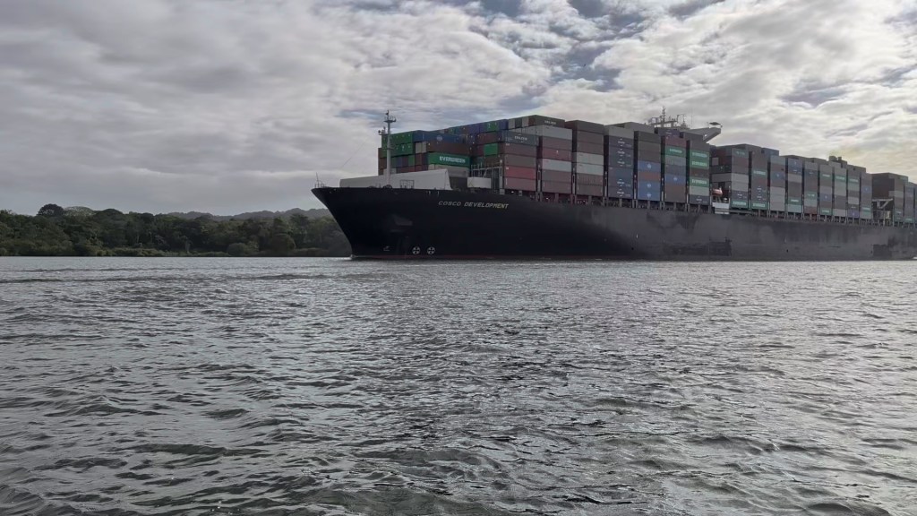 A large cargo ship with colorful containers on deck navigates a calm body of water under a cloudy sky.