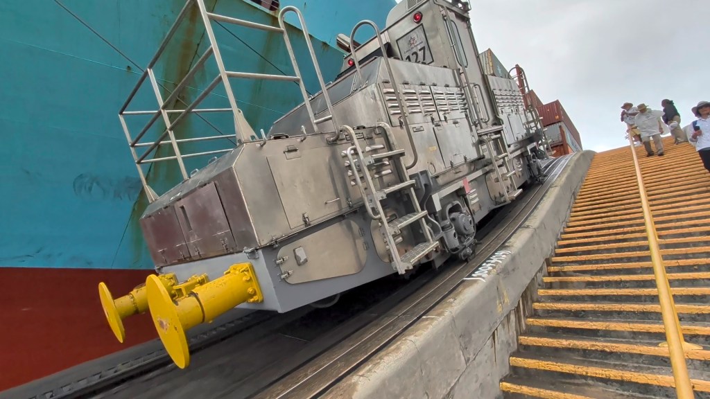 A train-like vehicle on a sloped docking ramp, unloading containers at a shipping terminal, with people walking nearby.