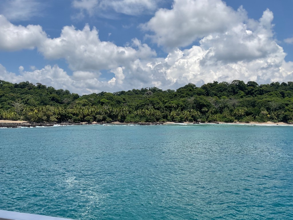 A scenic view of a tropical beach with lush green hills in the background, clear turquoise water, and a partly cloudy sky.