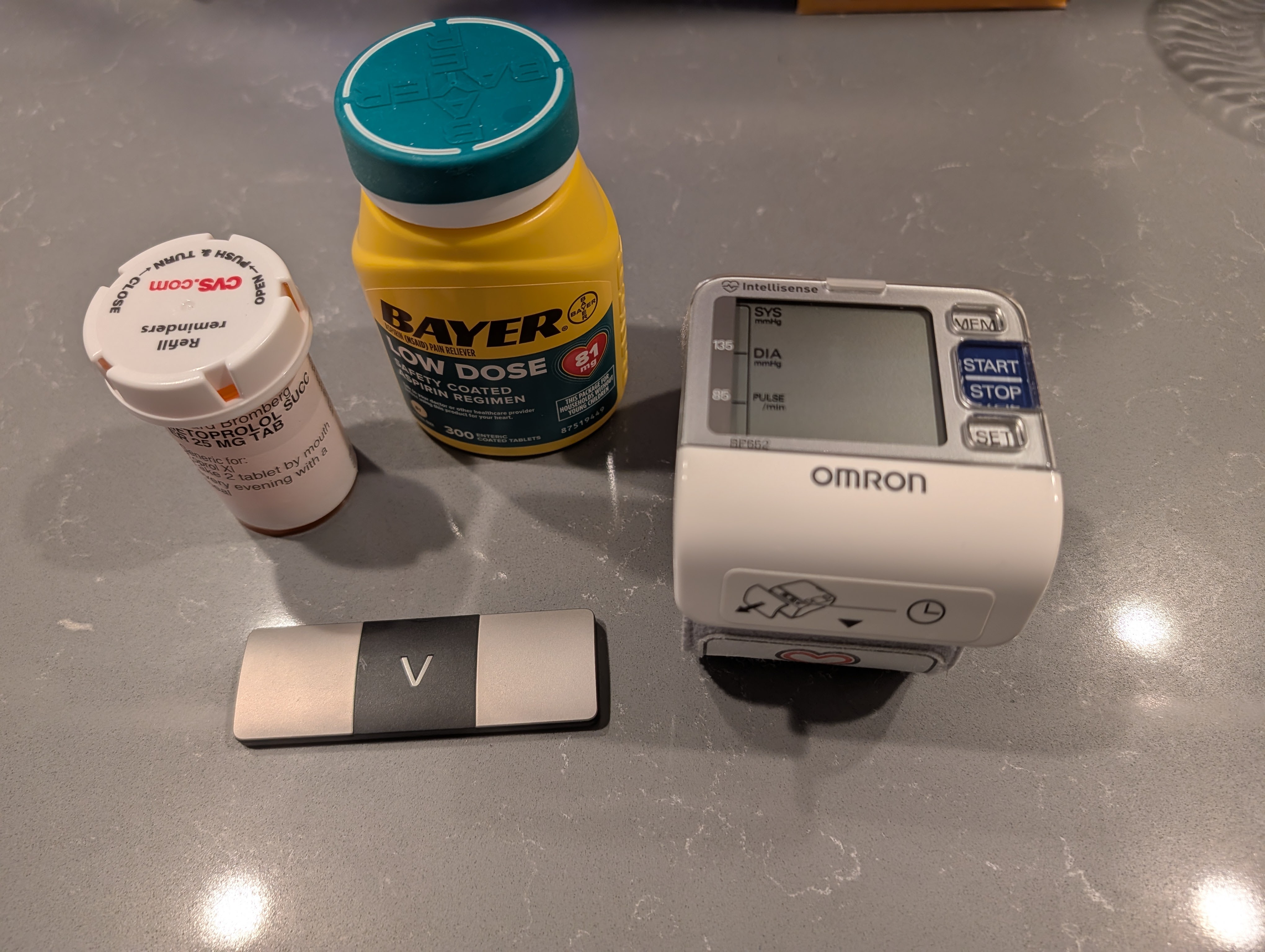 A blood pressure monitor, a bottle of Bayer low dose aspirin, a prescription medication bottle, and a small black and silver object arranged on a countertop.