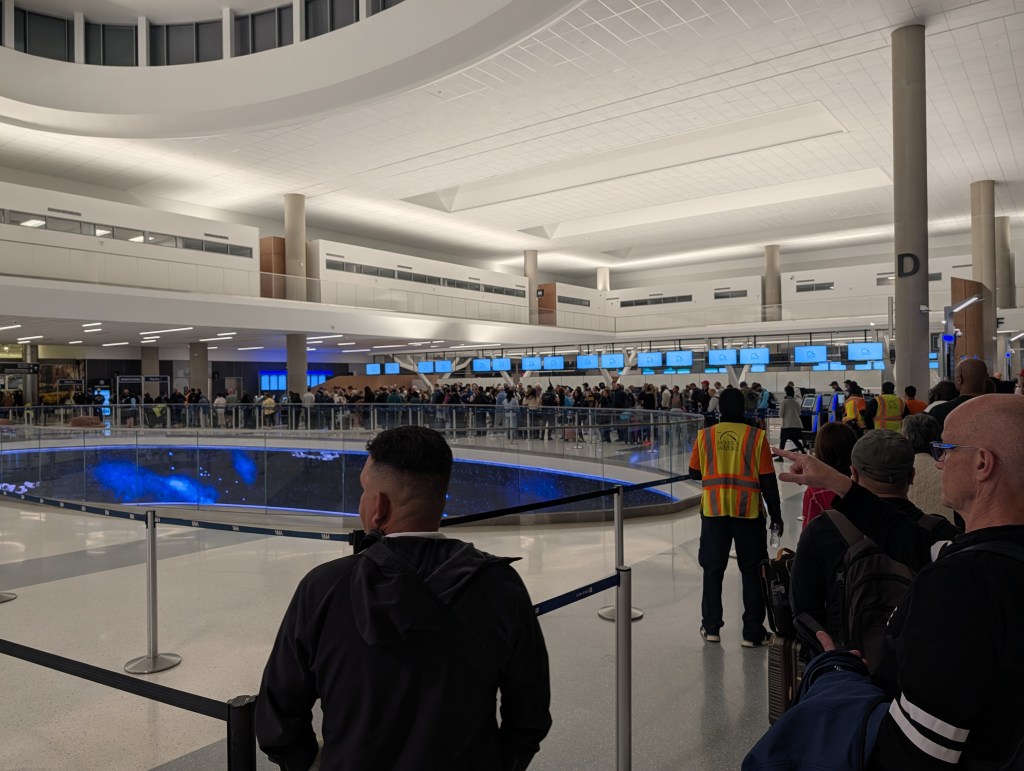 Busy airport terminal with a long check-in line, displaying multiple screens and travelers waiting.