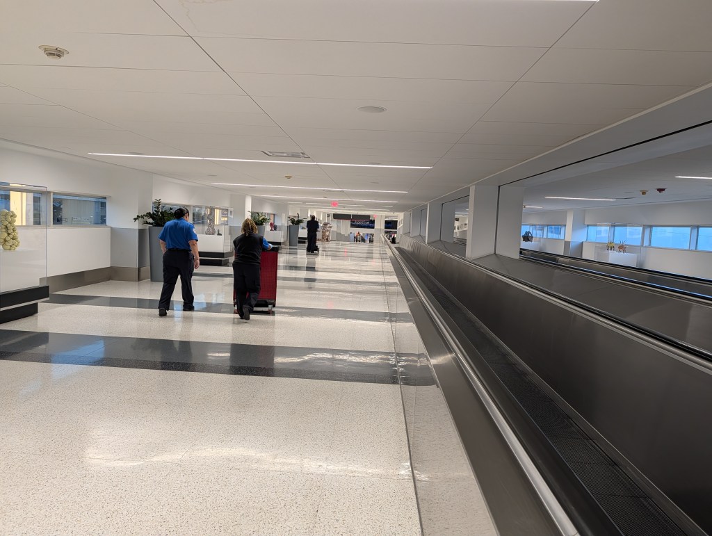 An airport hallway with travelers walking, one with a luggage cart. The area features bright lighting, polished floors, and large windows providing natural light.