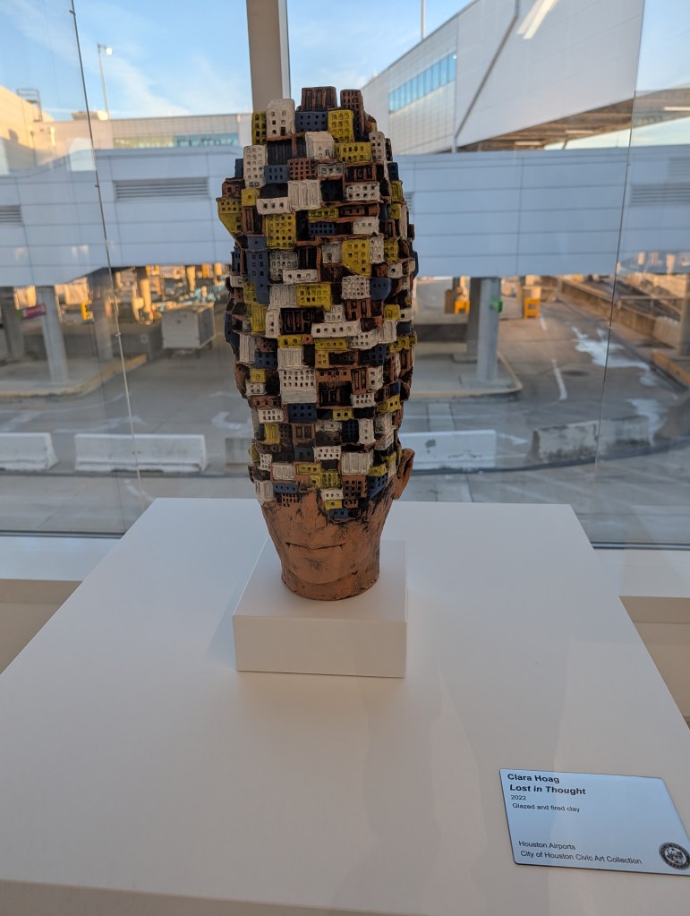 Sculpture titled 'Lost in Thought' by Clara Hoag, featuring a head made of glazed and fired clay, adorned with colorful painted brick-like structures, displayed in an airport setting.