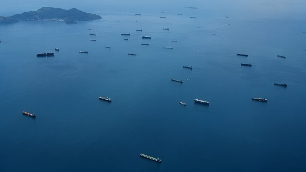 Aerial view of numerous cargo ships anchored in a calm sea, with an island in the background.