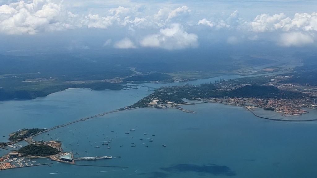 Aerial view of a coastal landscape featuring multiple waterways, boats in the water, and a town along the shore, with lush green hills and scattered clouds in the background.