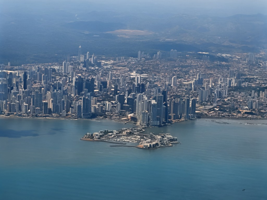 Aerial view of a coastal city featuring a skyline of tall buildings and a small island in the foreground with waterfront properties.