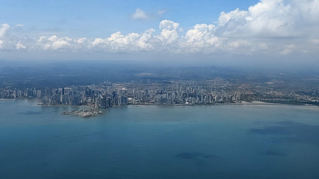 Aerial view of a coastal city with numerous high-rise buildings, surrounded by water and mountains under a blue sky with scattered clouds.