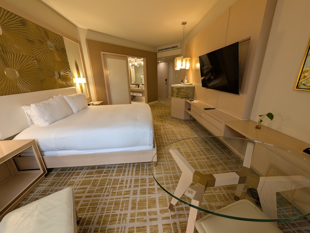 Modern hotel room featuring a queen bed, a glass-top dining table, and a flat-screen TV with contemporary decor and warm tones.