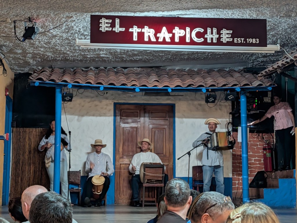 A live music performance at El Trapiche restaurant featuring musicians playing traditional instruments, with a sign displaying the restaurant's name and establishment year.