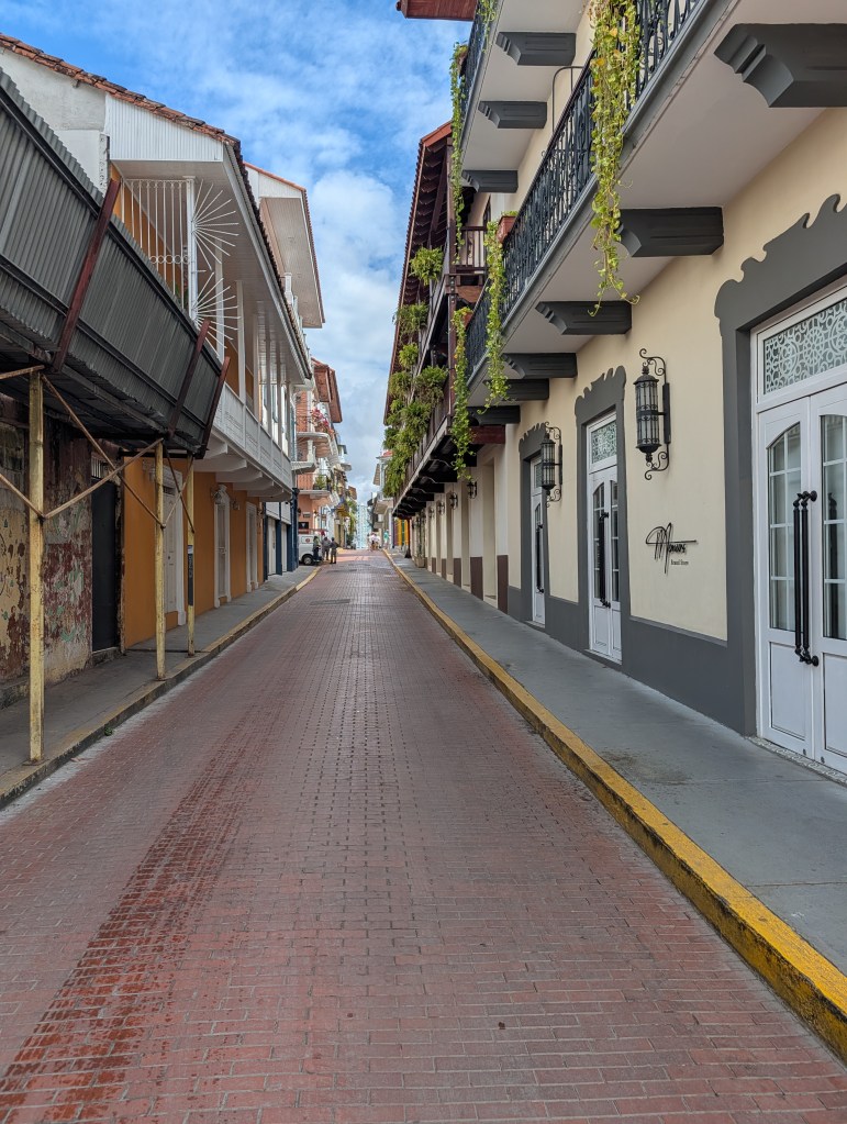 A narrow cobblestone street lined with colorful colonial buildings and balconies adorned with greenery, under a blue sky with scattered clouds.