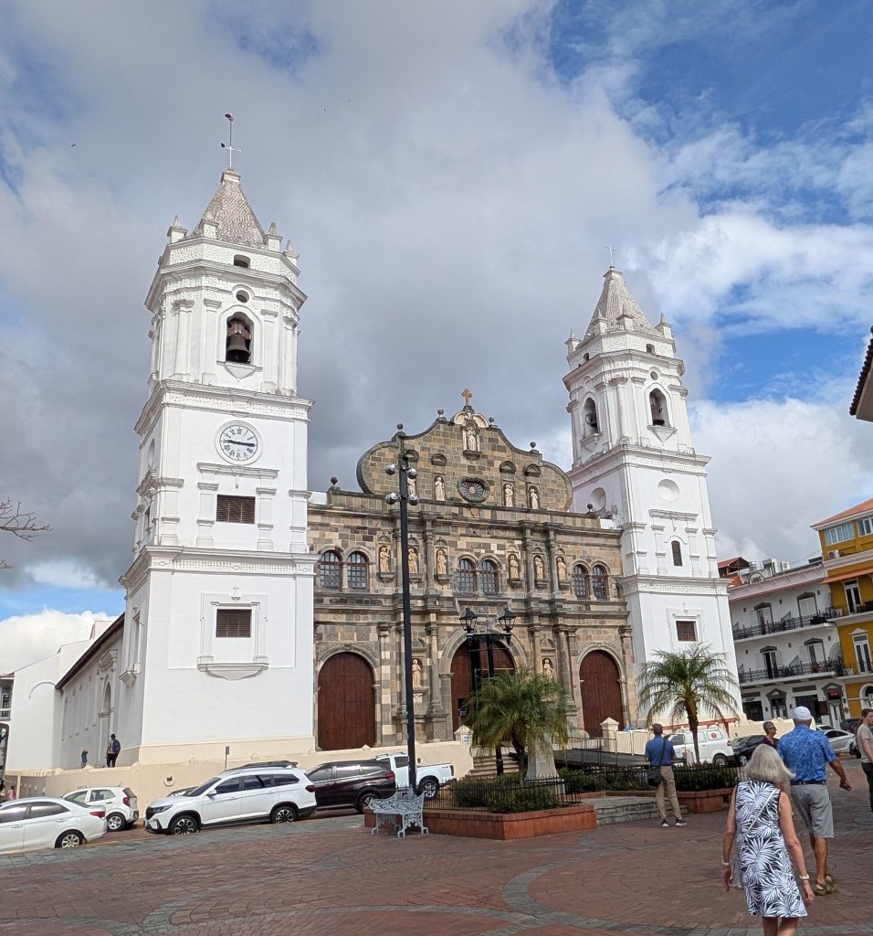 A historic church with two tall white towers and a detailed stone facade, set against a partly cloudy sky. People are walking in front of the church, and cars are parked nearby.