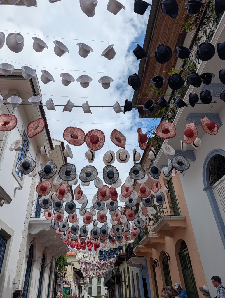 Colorful hats hanging overhead along a street, with buildings on either side and a cloudy sky above.
