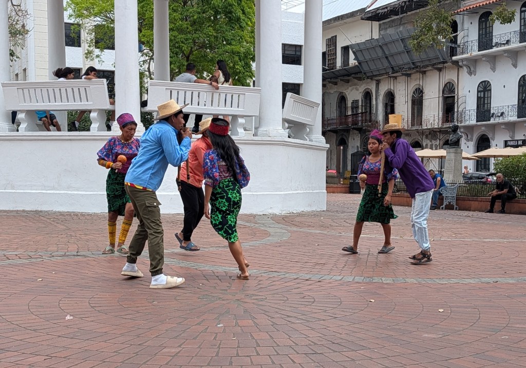 A lively outdoor scene featuring dancers in traditional colorful clothing performing in a plaza, surrounded by onlookers and colonial-style buildings.