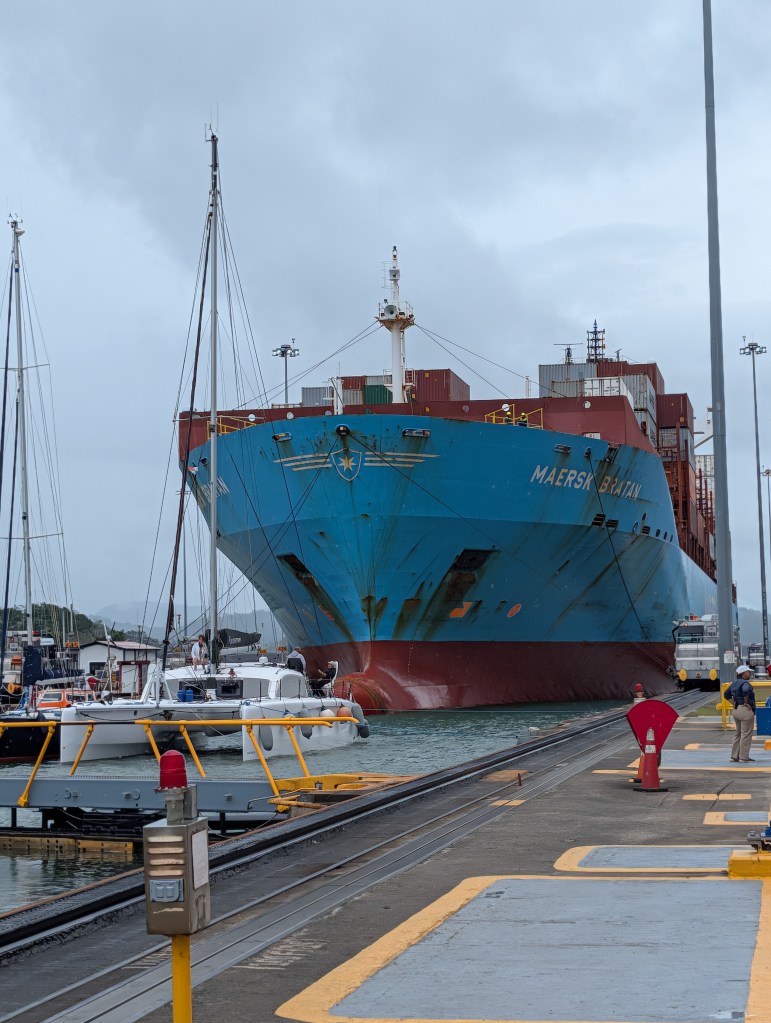 A large blue cargo ship, Maersk Batan, docked at a harbor with several smaller boats alongside. The scene includes cloudy skies and a port environment with a person standing on the dock.
