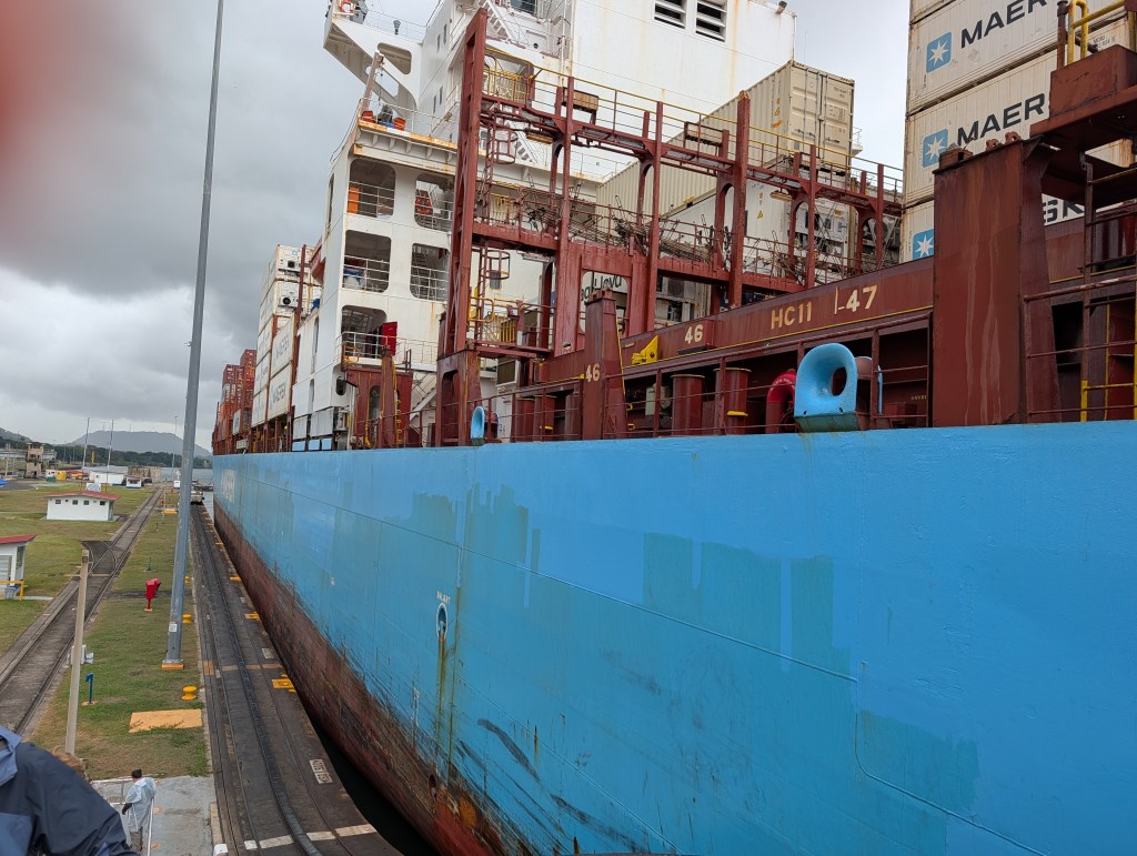 A large cargo ship passing through a canal, with visible shipping containers stacked on its deck, under a cloudy sky.