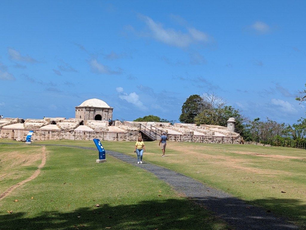 Two people walking on a path in front of an ancient fortification under a blue sky with a few clouds.
