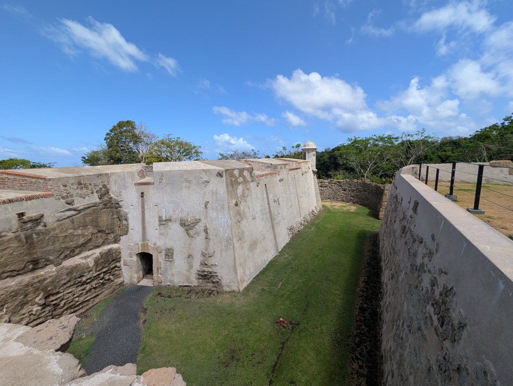 View of a historical fort with stone walls, a grassy area, and clear blue skies with scattered clouds.