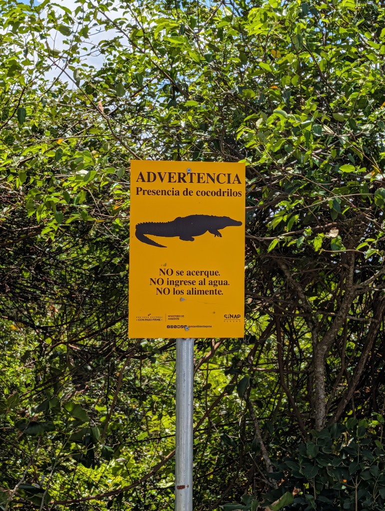Warning sign indicating the presence of crocodiles, with instructions not to approach, enter the water, or feed them, surrounded by greenery.