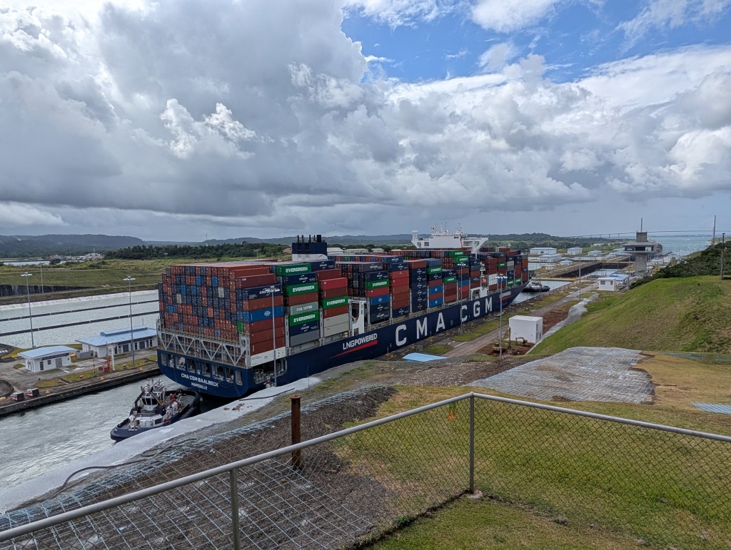 A large container ship, labeled CMA CGM, navigates through a canal with colorful shipping containers stacked on its deck, under a cloudy sky.