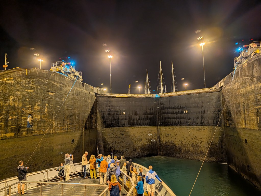 A group of people on a boat in a lock at night, with the lock gates partially open and illuminated by overhead lights.