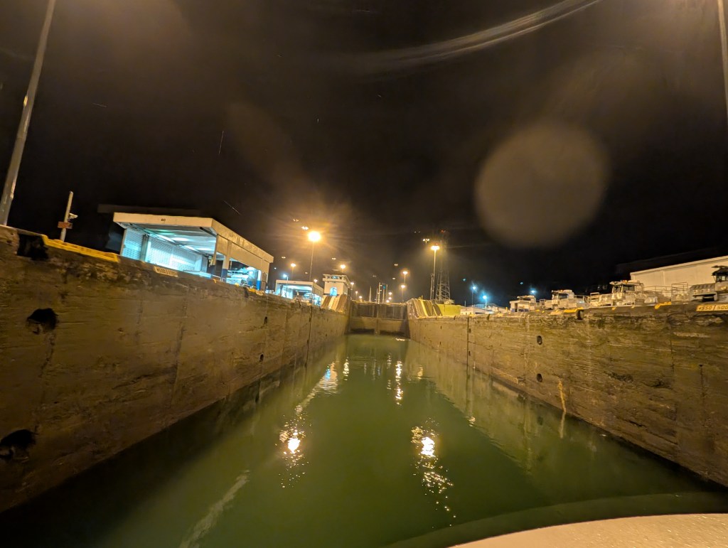 View of a canal lock at night, with illuminated buildings and boats on either side, reflecting in the water.