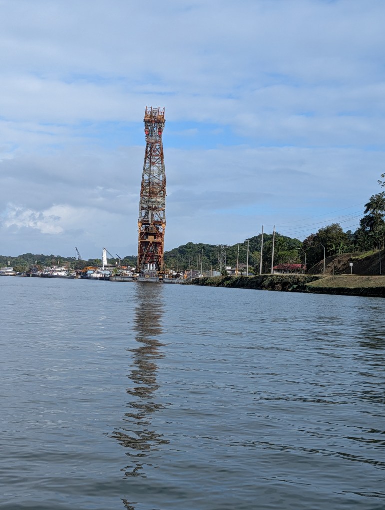 A tall industrial structure reflecting in calm water, surrounded by greenery and a cloudy sky.