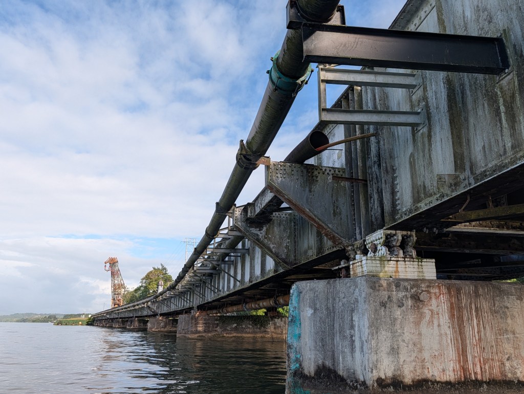Low-angle view of an industrial bridge with pipes running along the structure, reflecting in the water below, under a partly cloudy sky.