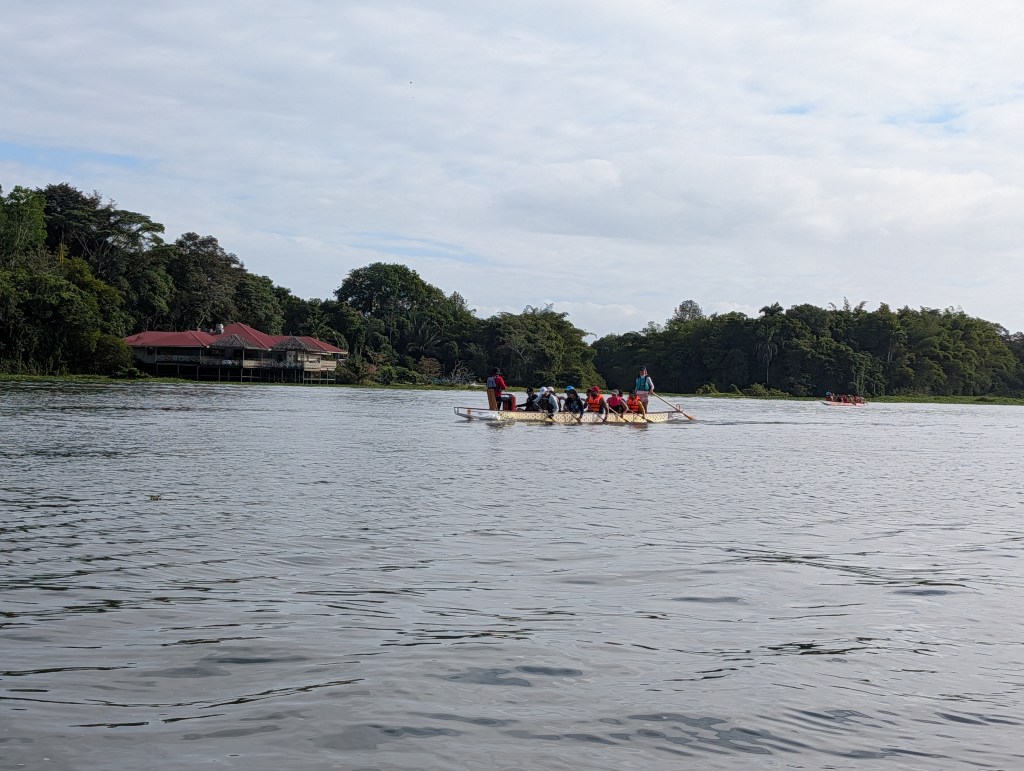 A group of people paddling in a boat on a calm river with green trees lining the shore and a building in the background.
