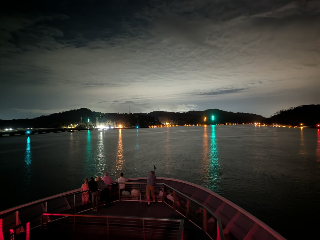 A view from the deck of a boat at night, showing a calm body of water reflecting colorful lights from nearby hills and a pier, with several people standing on the boat looking out.