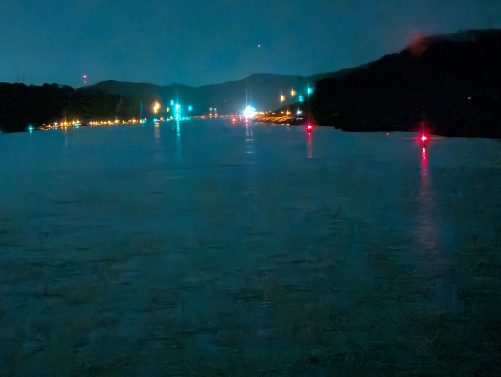 A nighttime view of a calm river or lake, illuminated by distant lights on the shoreline and reflecting colors on the water's surface.