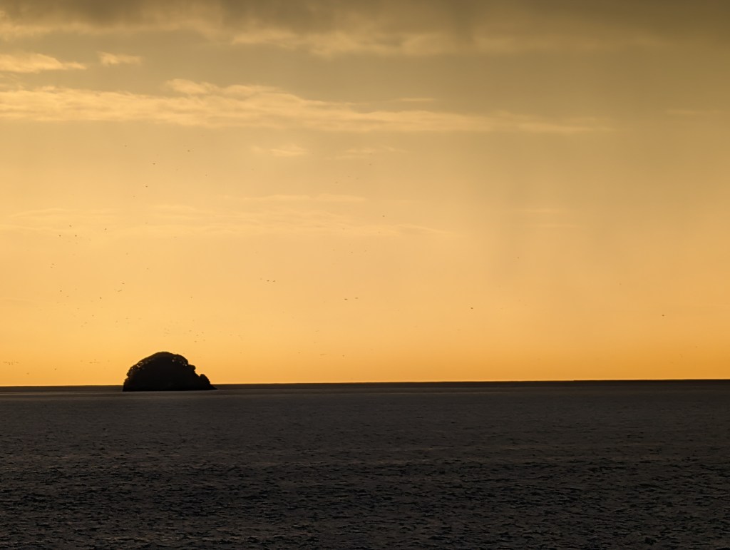 A distant rock formation silhouetted against a gradient sunset sky with hues of orange and yellow, above calm ocean waters.