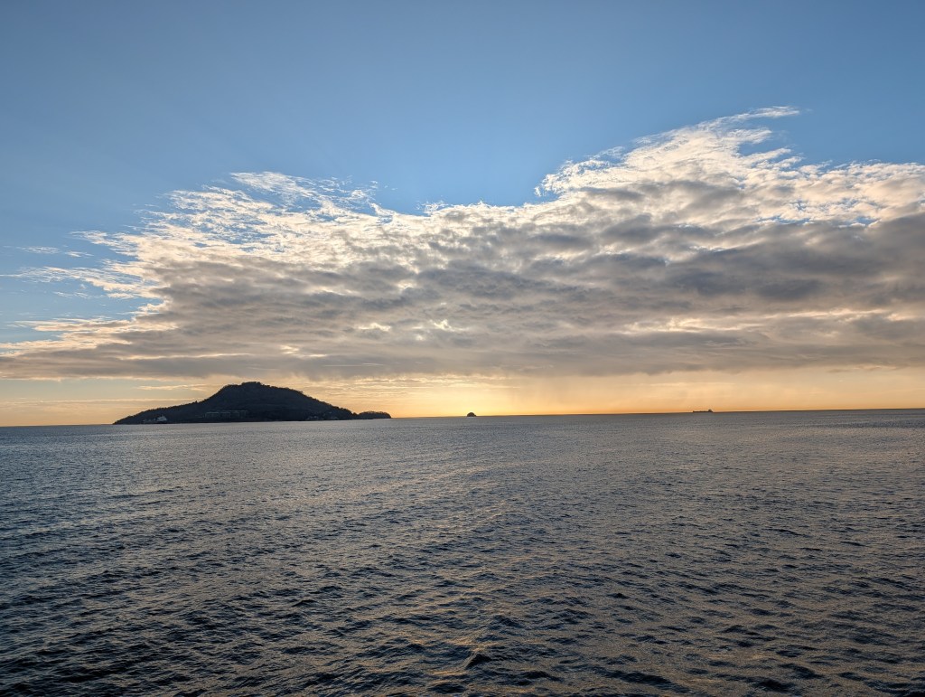 A panoramic view of a calm sea during sunset, featuring a distant island with a mountainous silhouette and scattered clouds in the sky.