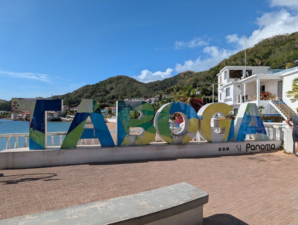 Colorful 'TABOGA' sign by the waterfront in Taboga, Panama, with mountains and buildings in the background.