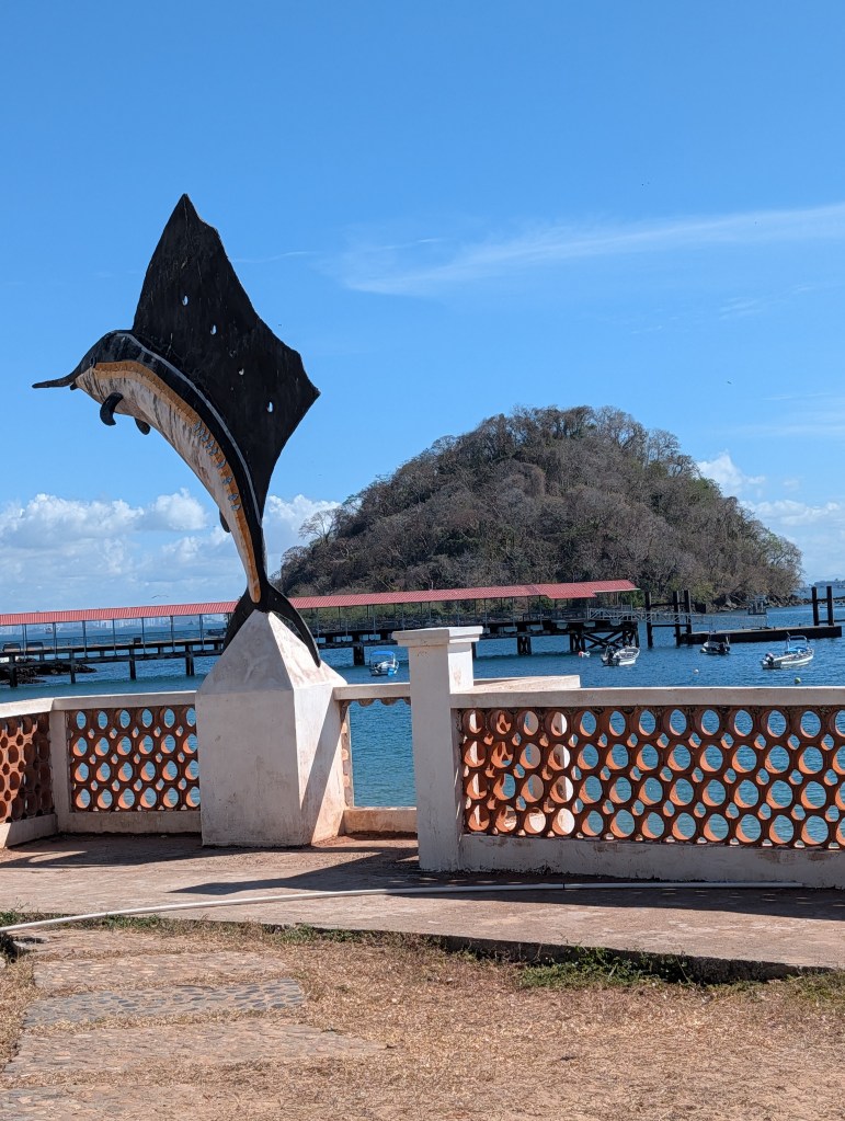 A large fish sculpture stands at the edge of a waterfront, overlooking a calm blue sea and a small island in the background. A wooden pier is visible with several boats moored nearby, under a clear sky.