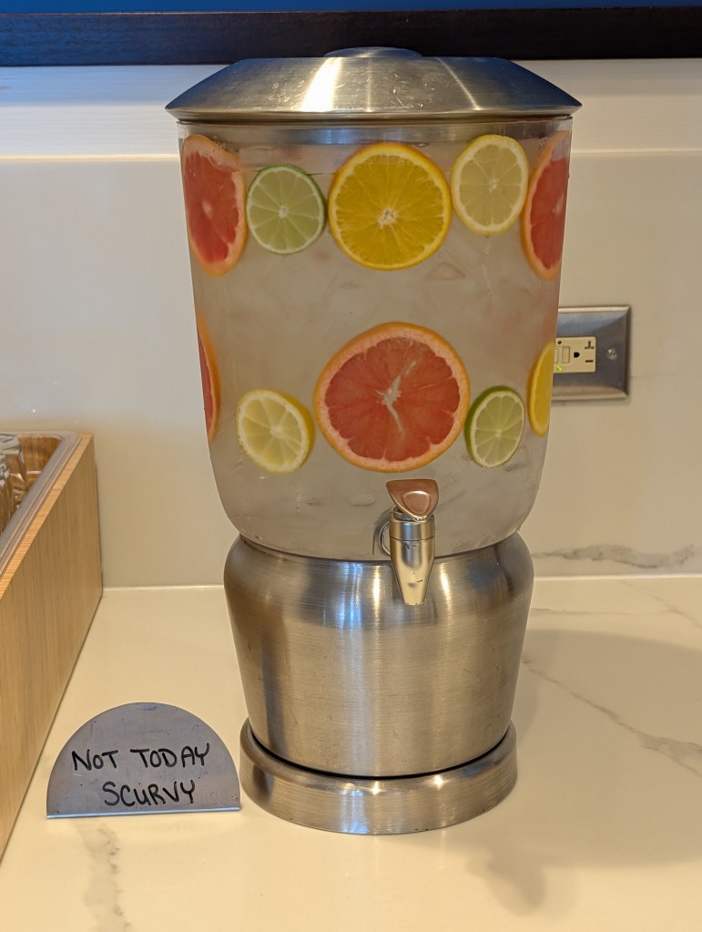 A beverage dispenser filled with water and slices of citrus fruits, including grapefruit, lemon, lime, and orange. A sign nearby reads 'Not Today Scurvy.'