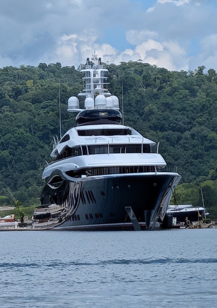 A large luxury yacht anchored near a lush green hillside, with a clear blue sky and clouds in the background.