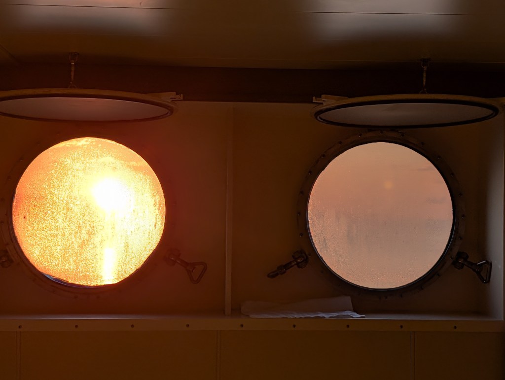 Two round portholes on a ship, one displaying a warm sunset glow and the other showing a foggy exterior view.