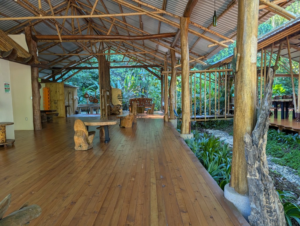 Interior view of a wooden structure surrounded by greenery, featuring bamboo columns, wooden furniture, and a spacious wooden floor.