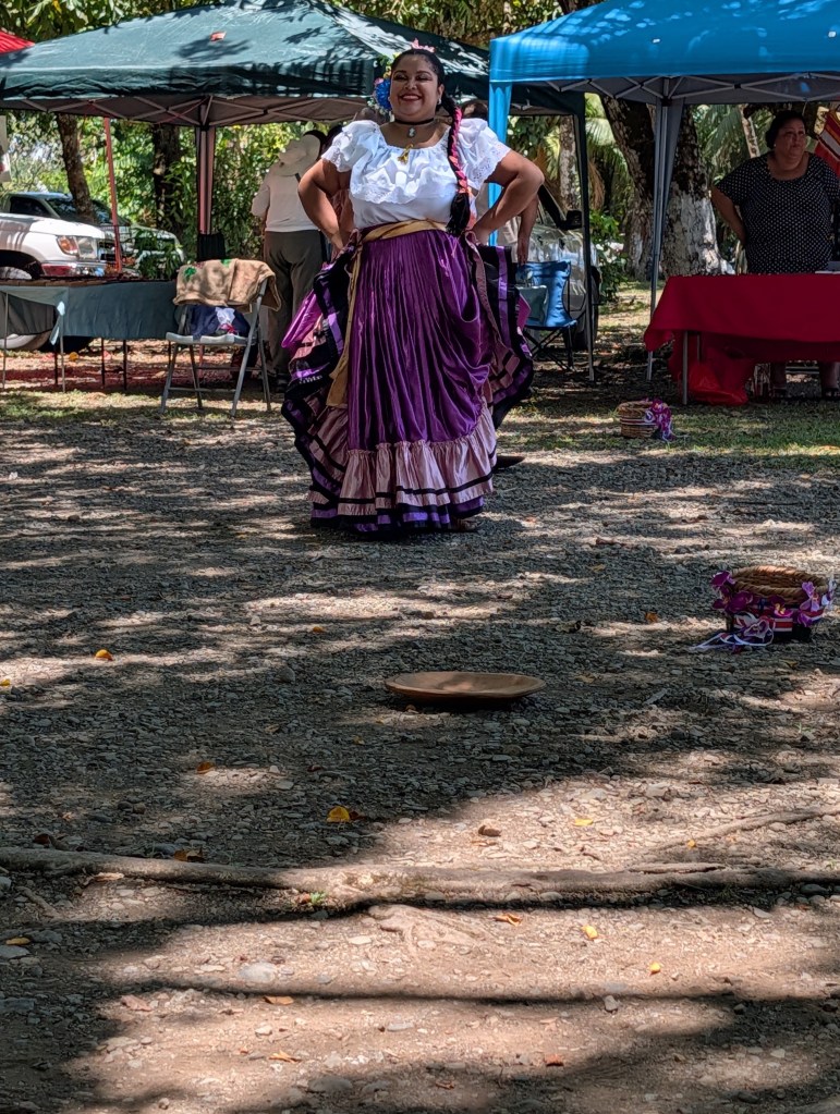 A woman dressed in traditional attire with a white blouse and a purple skirt is dancing outdoors. The setting includes canopies and other people in the background, creating a festive atmosphere.