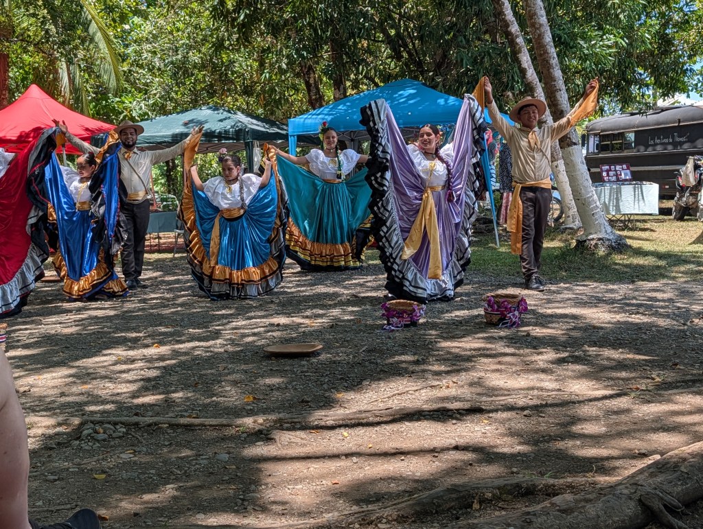 A group of dancers performing a traditional dance outdoors, wearing colorful folkloric costumes. The scene features bright blue, red, and gold outfits, with dancers showcasing vibrant movements under sunny trees.