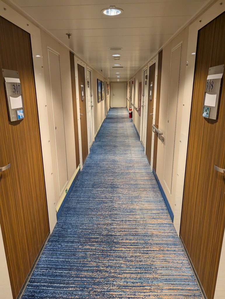 A long hotel hallway featuring numbered doors and carpeted flooring in blue tones, illuminated by ceiling lights.