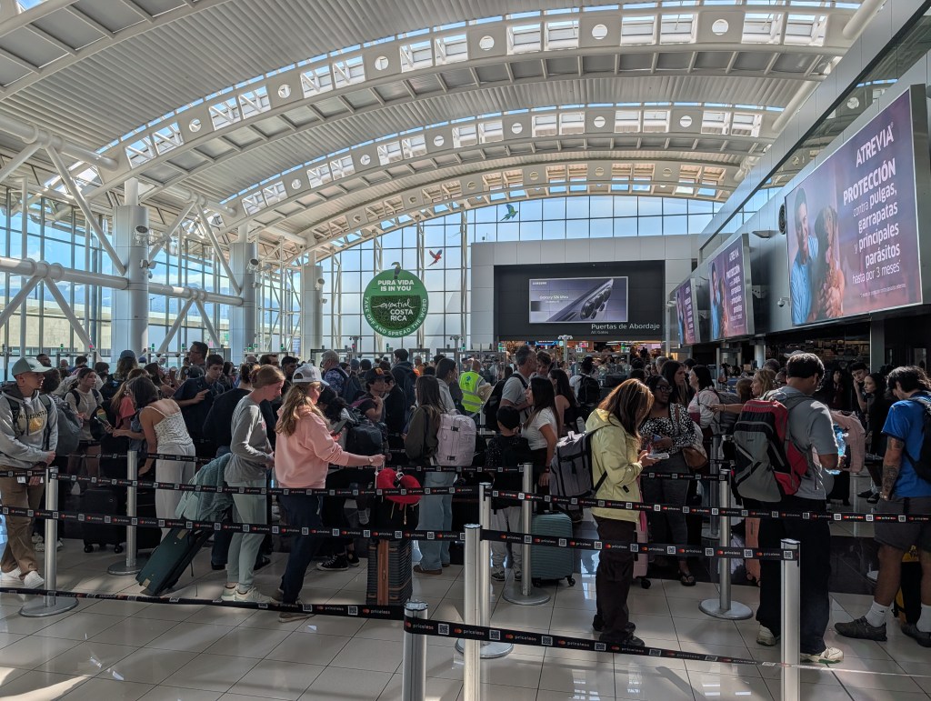 Crowded airport terminal with a long line of passengers waiting in front of check-in counters, large windows allowing natural light, and several advertisements visible in the background.