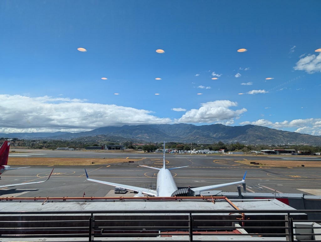 View from an airport window showcasing airplanes on the tarmac with mountains and blue sky in the background.