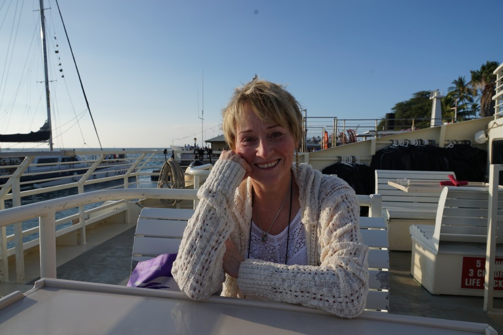 A woman sitting at a table on a boat deck, smiling at the camera with a serene ocean background and a clear blue sky.