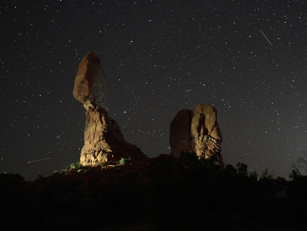 A nighttime landscape featuring distinctive rock formations under a starry sky, with a visible meteor streaking across the scene.
